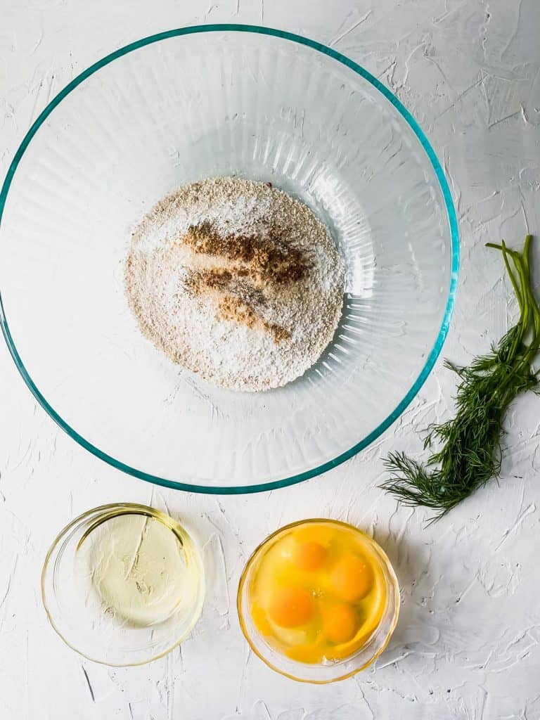 matzo ball dry ingredients in a glass bowl with eggs, oil, and dill on the side