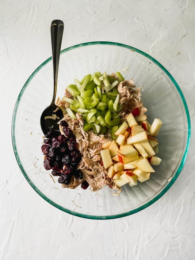 shredded chicken and diced apples and celery in a glass bowl with dried cranberries