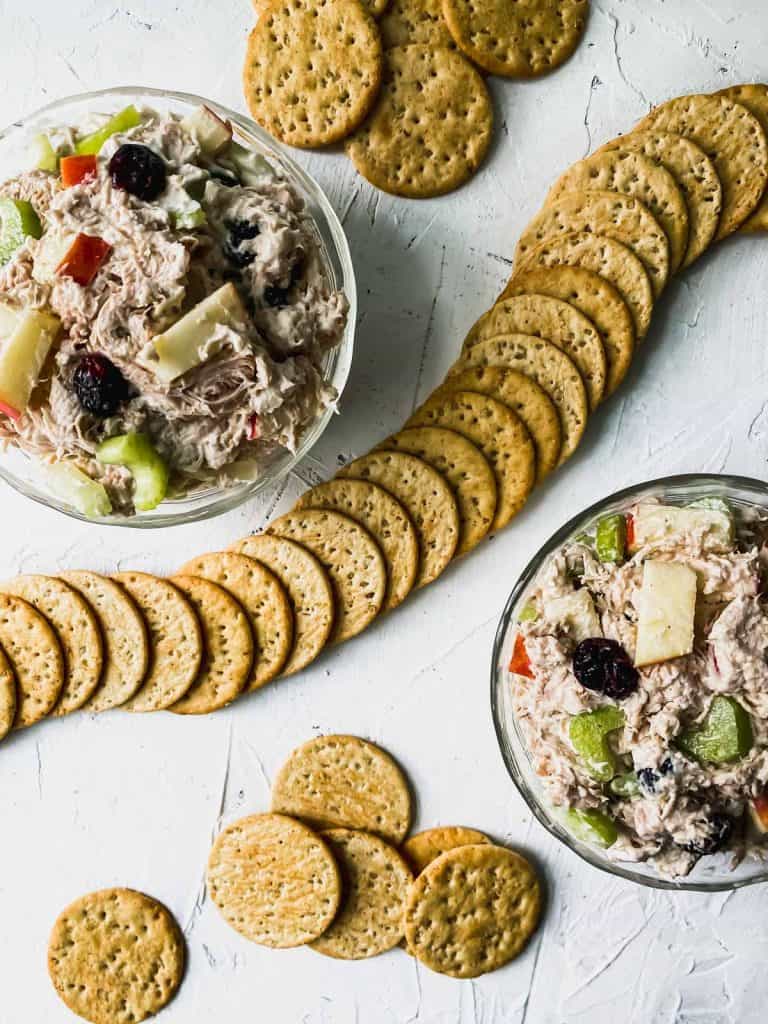 2 glass bowls of shredded chicken salad with cranberries and apples with crackers around them