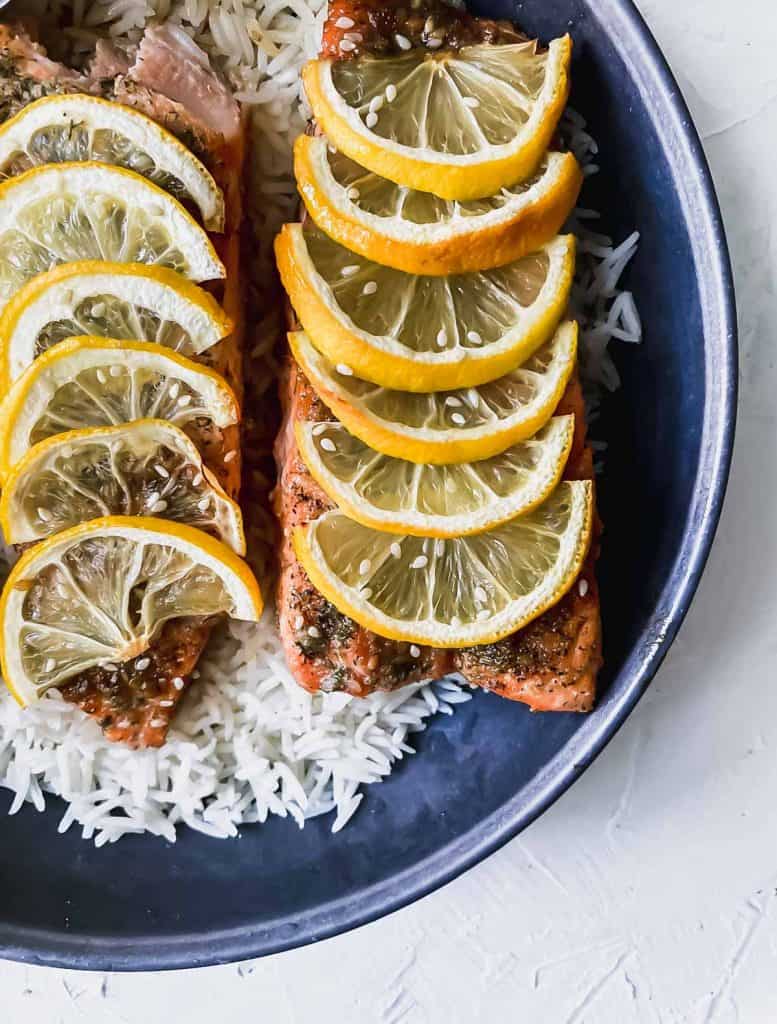 teriyaki salmon with lemon slices on top on a bed of rice in a black bowl.
