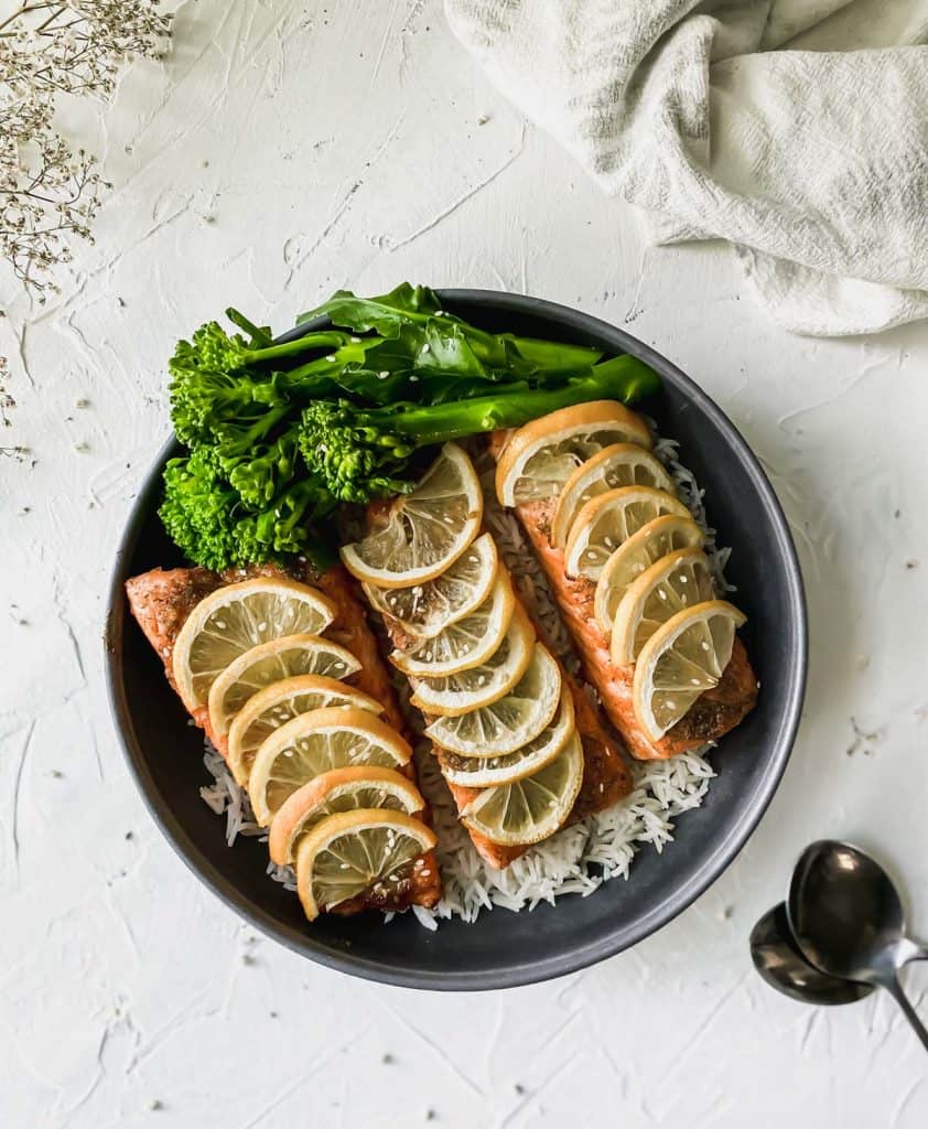 black bowl full of white rice, broccoli, and salmon with 2 spoons below it.