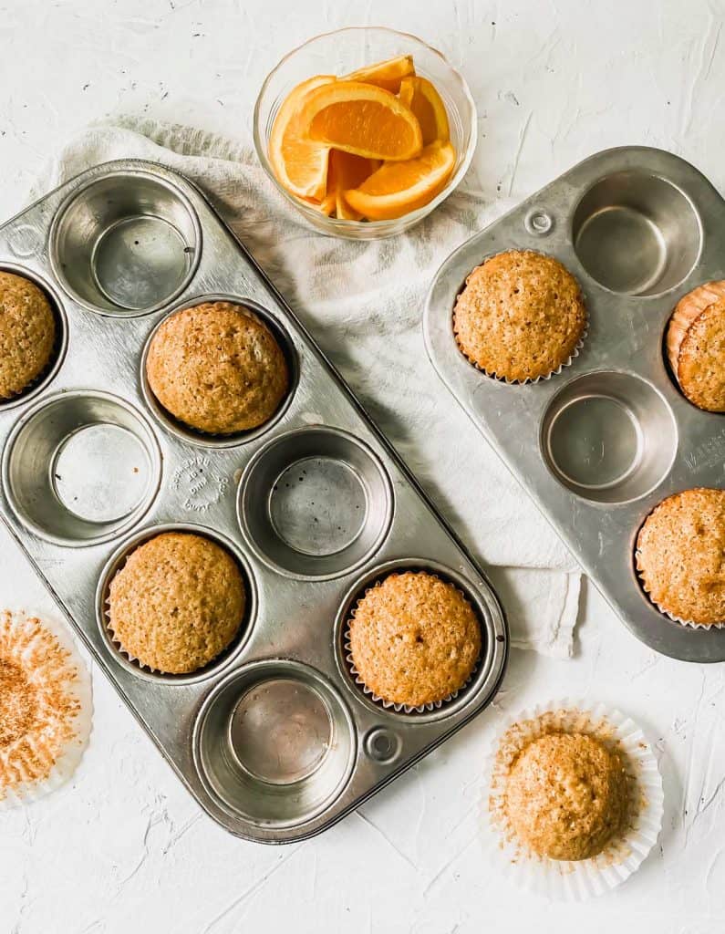 Spiced orange muffins in muffin pans with orange slices next to them.